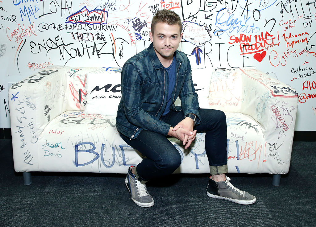 A man sits in a room full of graffiti wearing demin pants and a denim jacket.