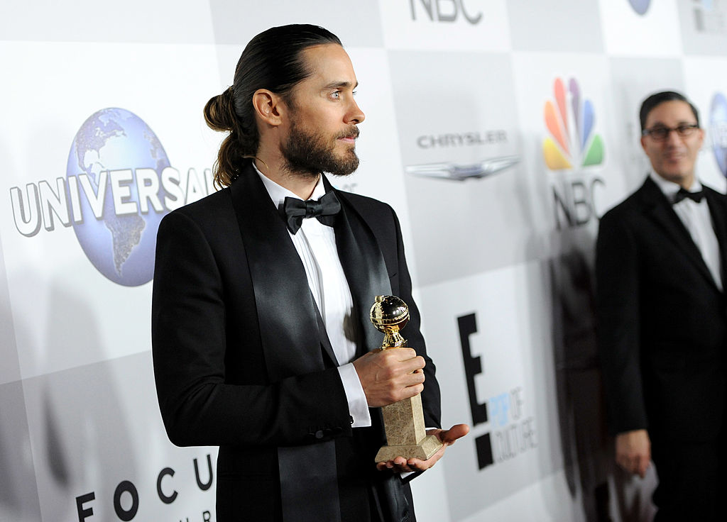 An actor and musician holds an award sporting a tuxedo and a man-bun.