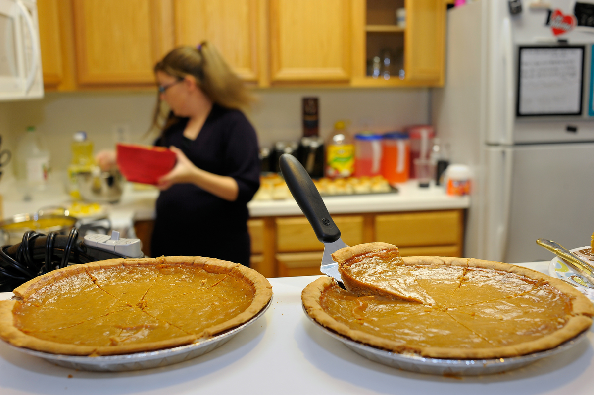 Two pumpkin pies sit on the kitchen counter as a woman prepares a dish.