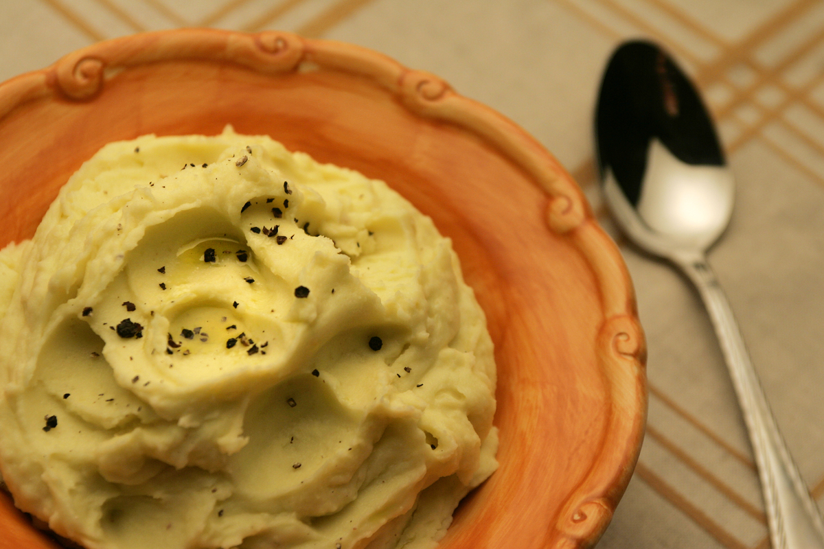 Mashed potatoes with olive oil are in an orange bowl with a spoon nearby.