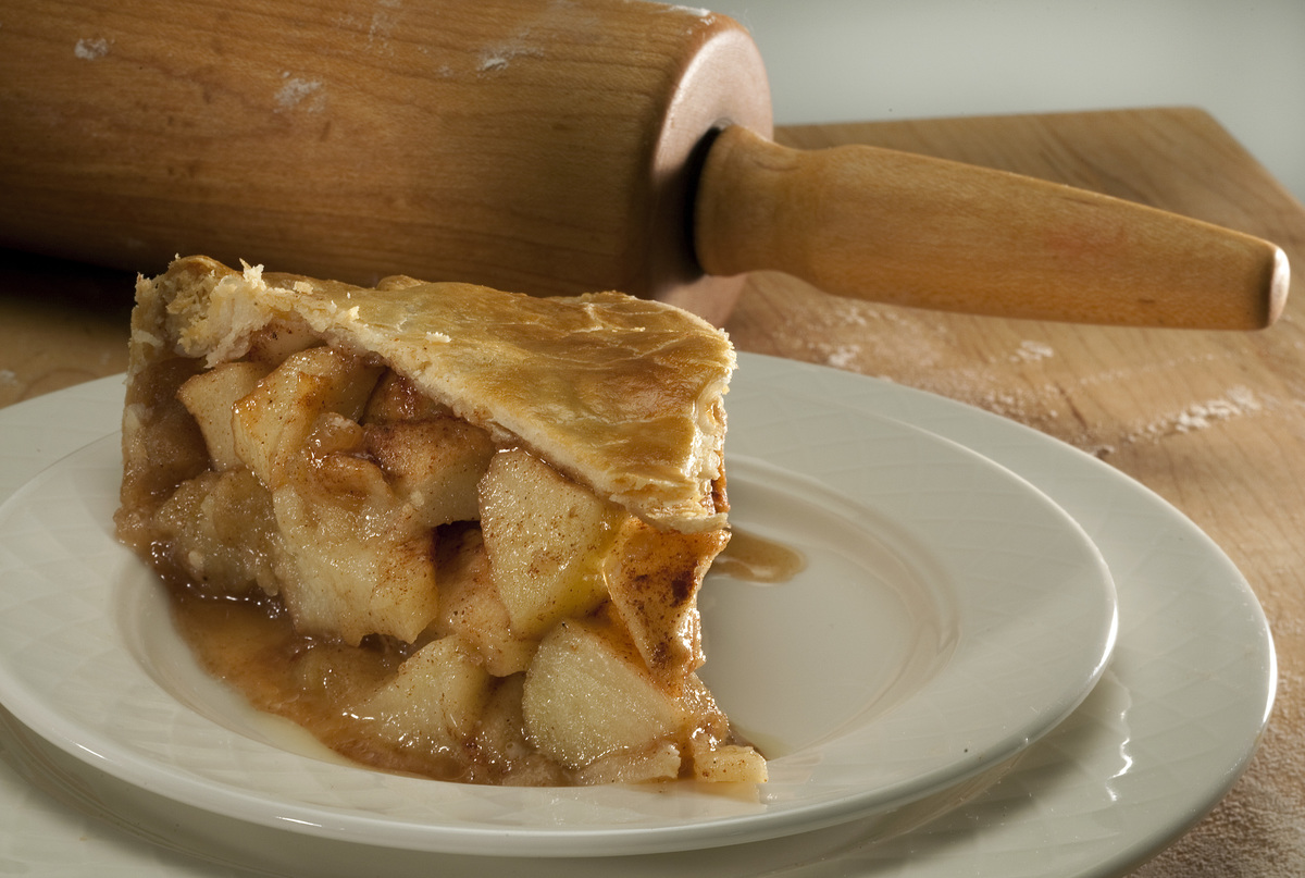 A slice of apple pie sits on a plate in front of a rolling pin.