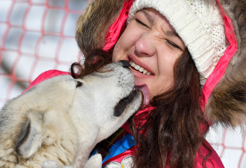 A woman squints while a dog licks her chin.