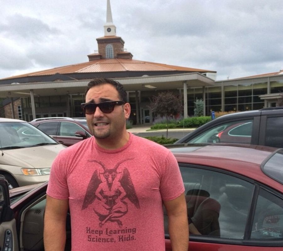 a guy wearing a tshirt that says 'keep learning science, kids' with a demon on it, he's standing in front of a church