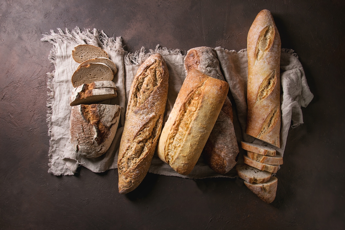 Variety of loafs fresh baked artisan rye, white and whole grain bread on linen cloth over dark brown texture background Top view, copy space