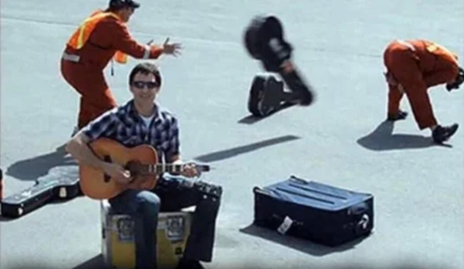 man with guitar next to airport workers throwing a guitar case