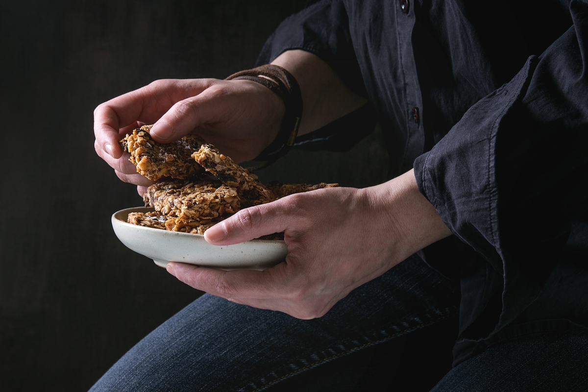 Woman in black shirt holding ceramic plate with homemade energy oats granola bars with dried fruits and nuts over black background. Healthy snack.