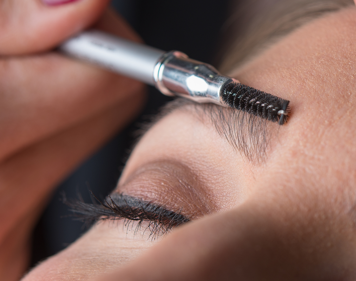 A makeup artist straightens a woman's eyebrows with a small brush.