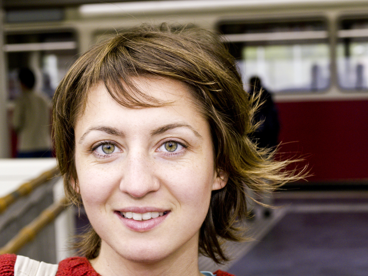 A young woman with green eyes smiles in front of a subway.
