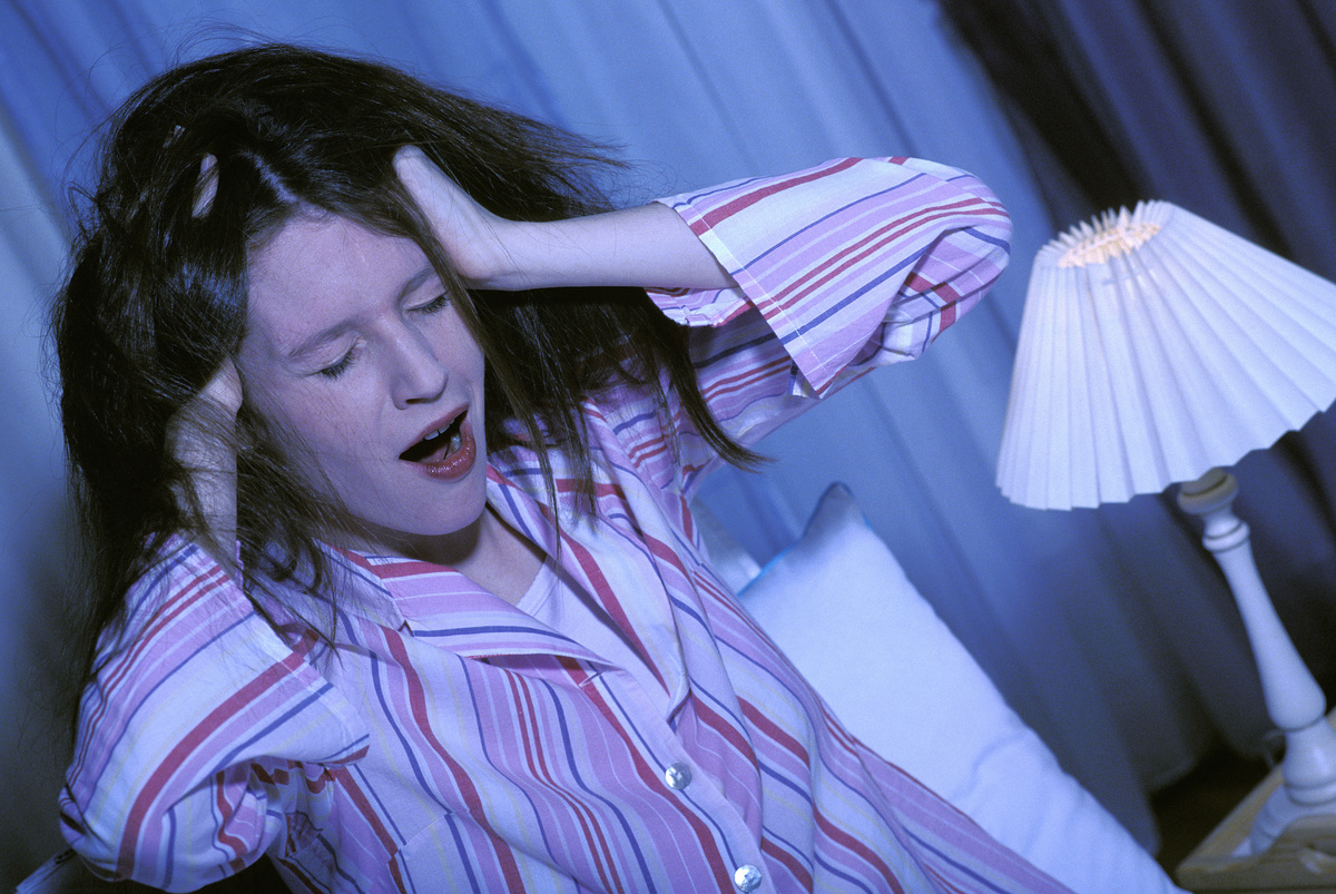 A woman with messy hair yawns while sitting up in bed.