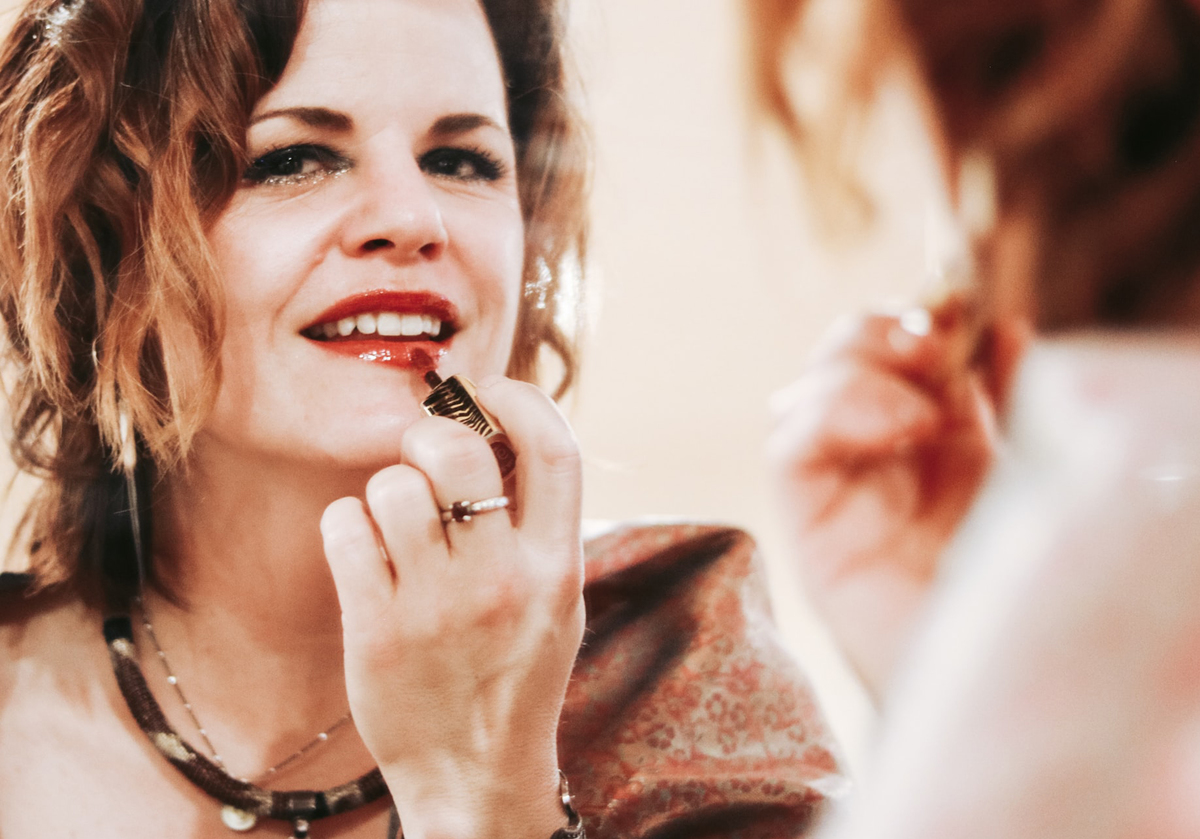 A woman applies red lipstick while looking in a mirror.