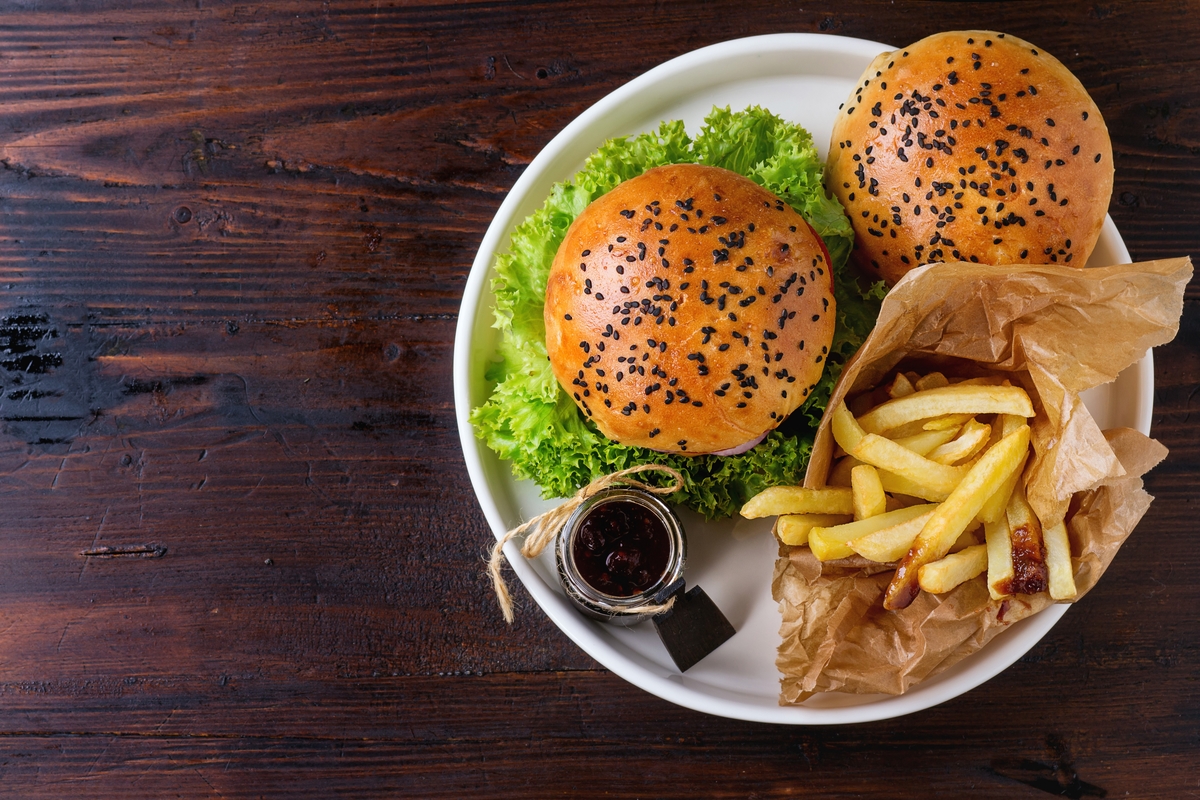 Fresh homemade burger with black sesame seeds in white plate with fried potatoes, served with ketchup sauce in glass jar over dark wooden table. Top view