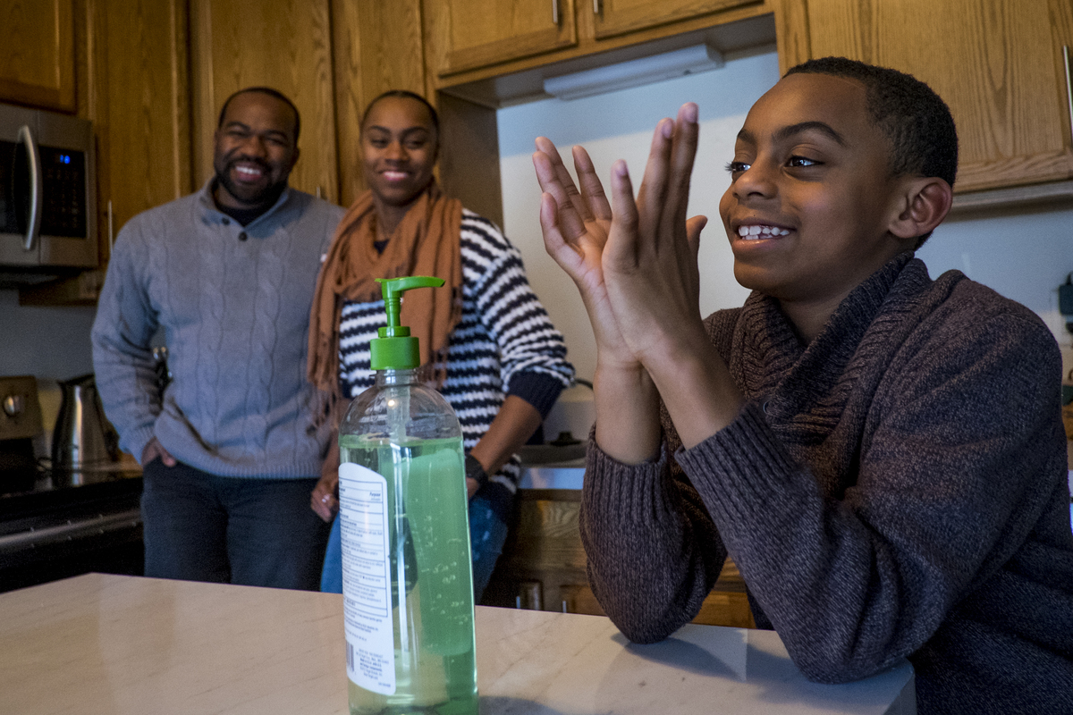 A boy uses hand sanitizer at a kitchen table as his parents smile in the background.