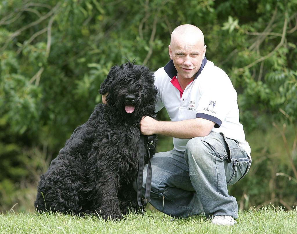A Black Russian Terrier sits in the grass next to its owner.
