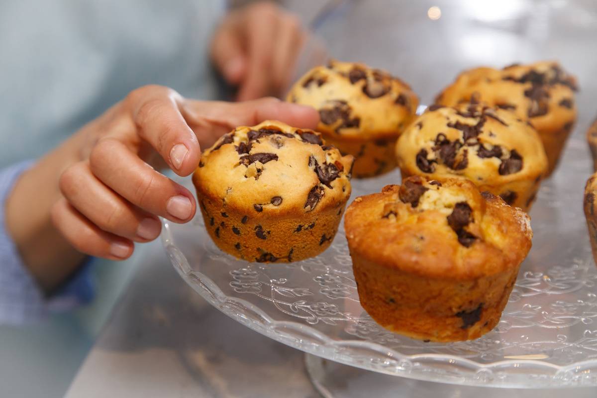 A woman selects a chocolate muffin from a tray.