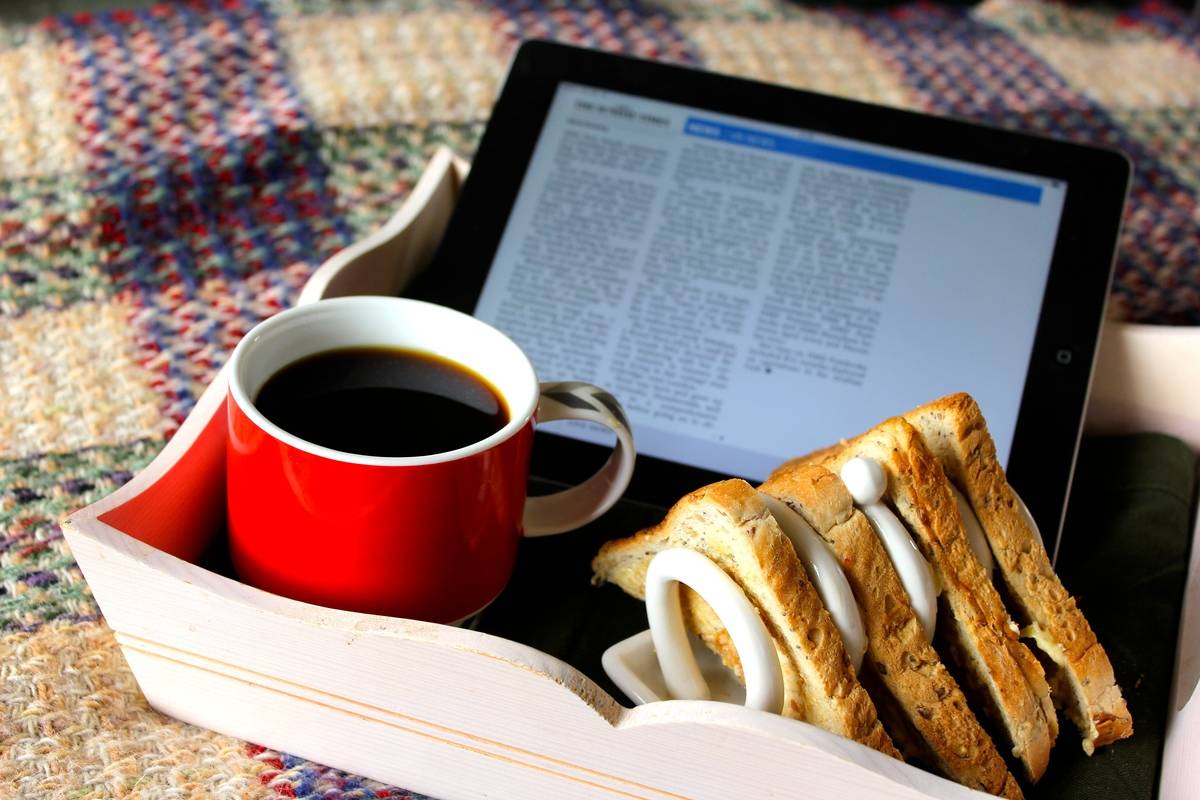 A breakfast tray features coffee, a sandwich, and the newspaper on an iPad.