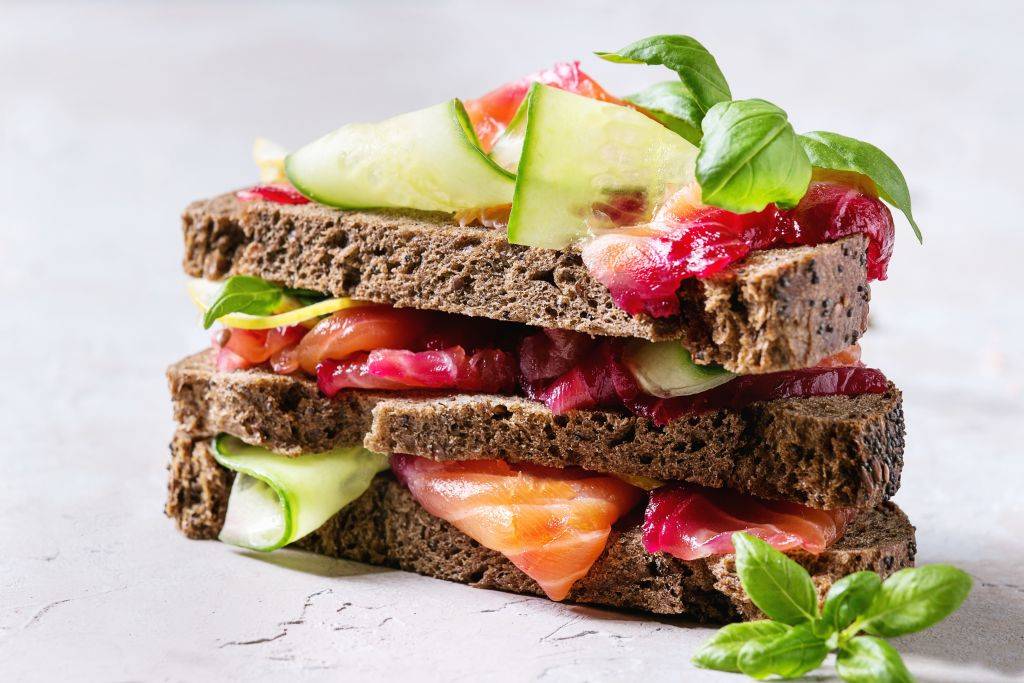 Sliced beetroot marinated salmon sandwiches with rye bread, cucumber, basil and lemon served in stack on wooden cutting board over grey blue texture background