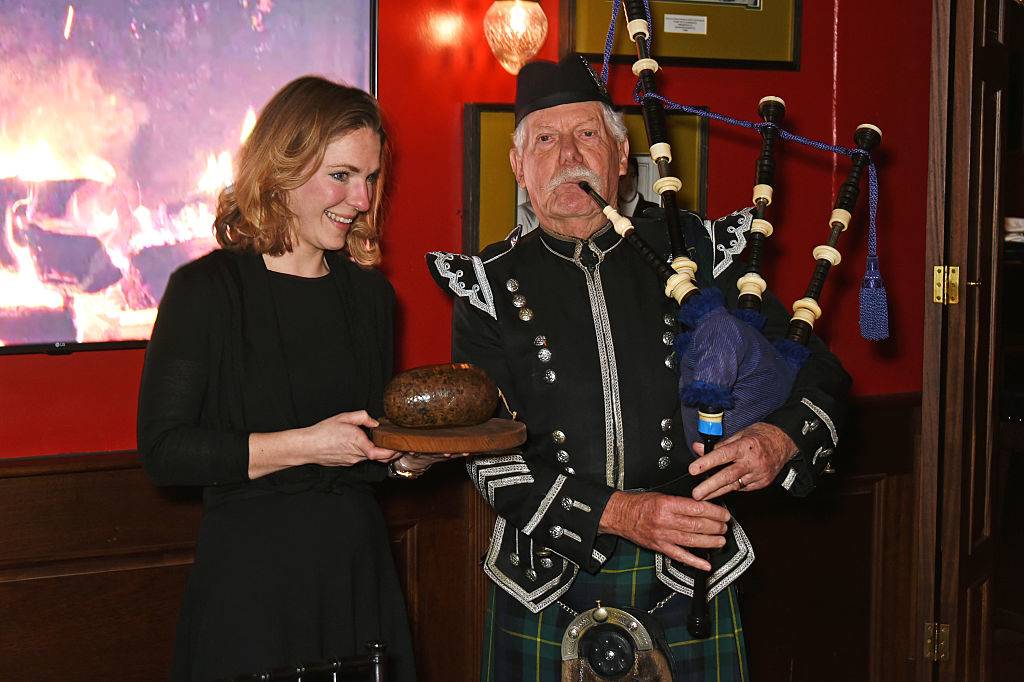 a woman holding haggis next to an old man playing a bagpipe in a kilt