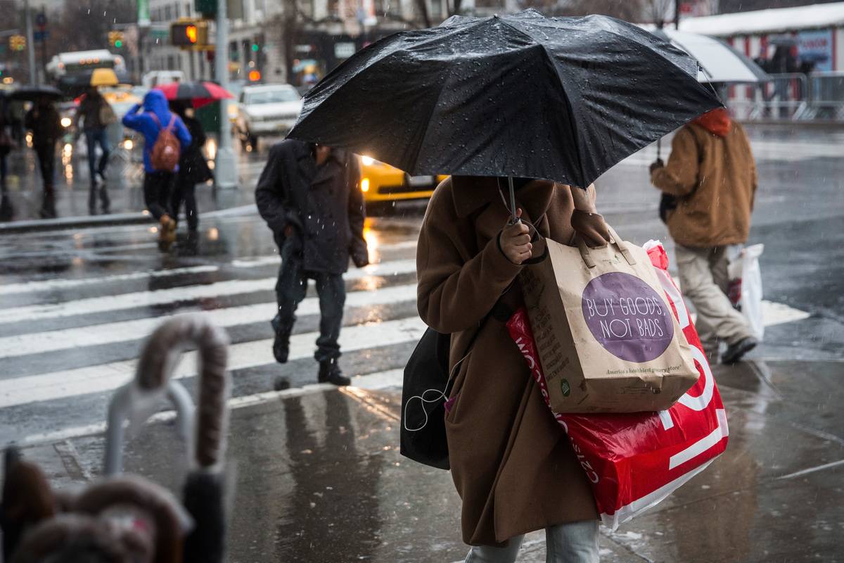 People walk along the sidewalk in the rain holding shopping bags