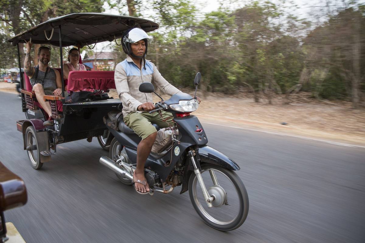 A Cambodian man drives two smiling Western tourists on his a motorbike Tuk Tuk