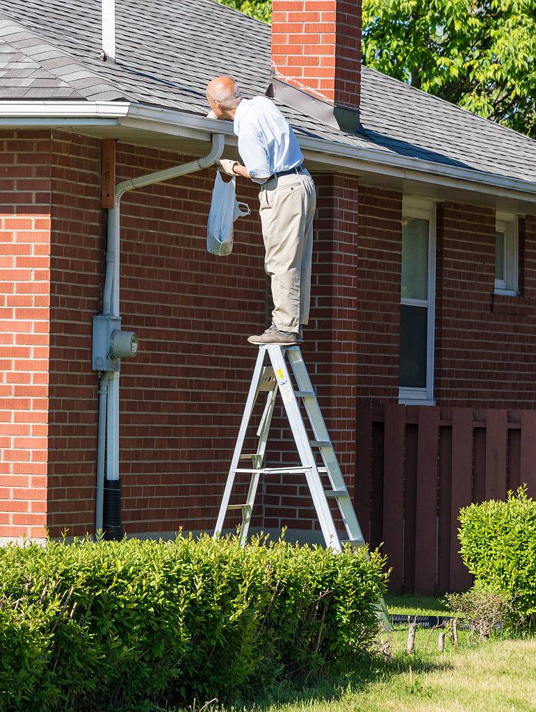 Seniors in Canada: Senior man cleaning a rain gutter on a ladder. Clearing autumn gutter blocked with leaves by hand.