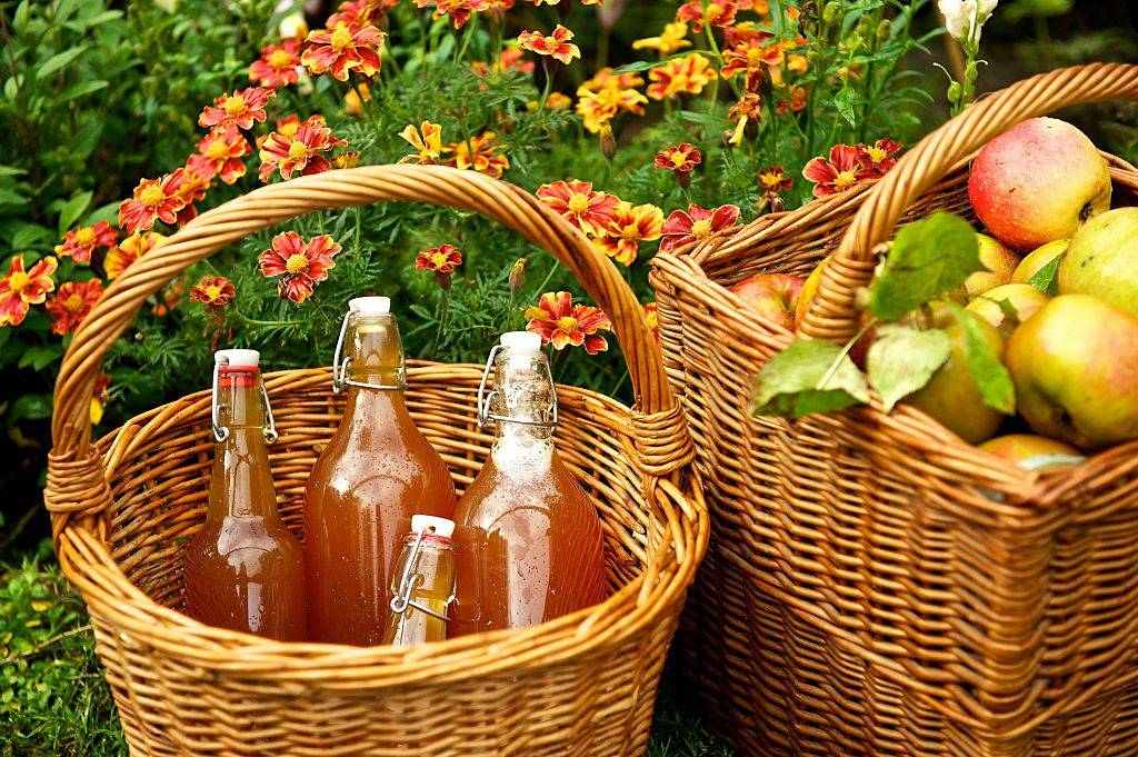 apple cider in a basket next to a basket of apples in front of flowers