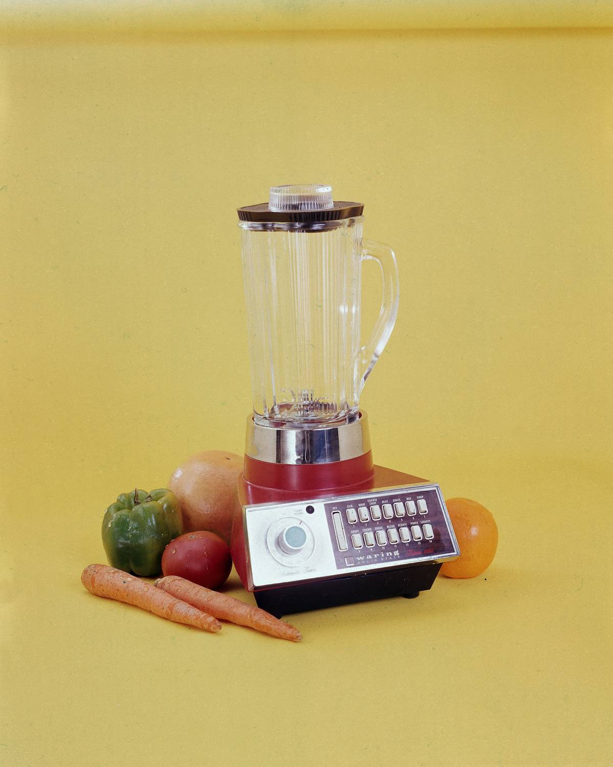 blender sits amid fruits and vegetables against a yellow background
