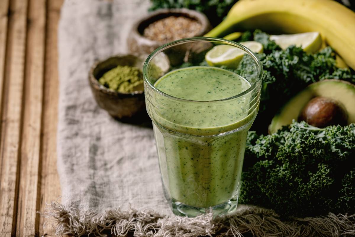 Glass of green healthy vegan smoothie. glass straw. Ingredients above. Kale. bananas. avocado. lime. non-diary milk. matcha powder and seeds over wooden plank cloth background. Close up