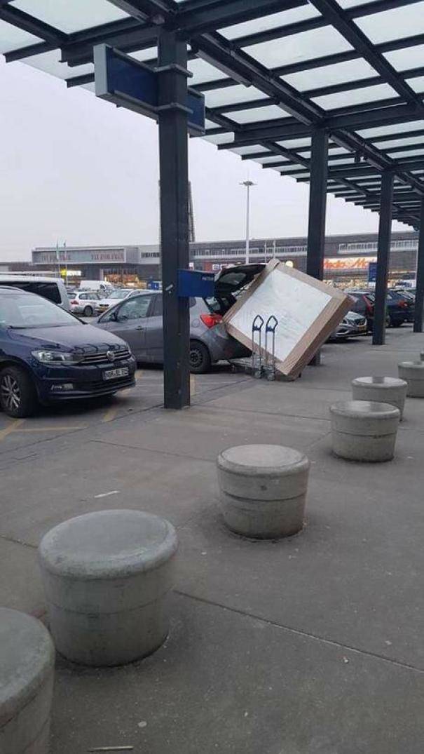 person trying to fit comically large box into the trunk of their car in a parking lot.