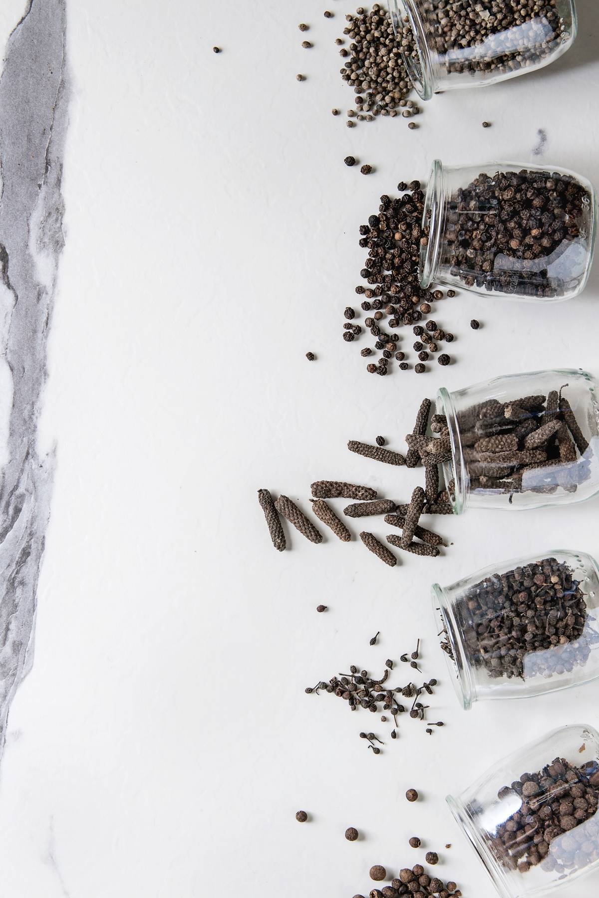 A variety of black peppers pour out of glass containers.