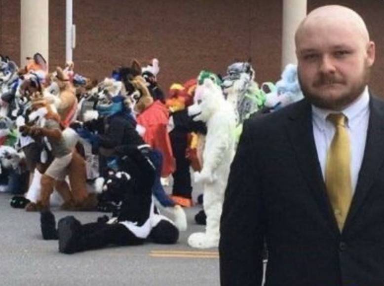 man in suit and tie with large group of furries in the background