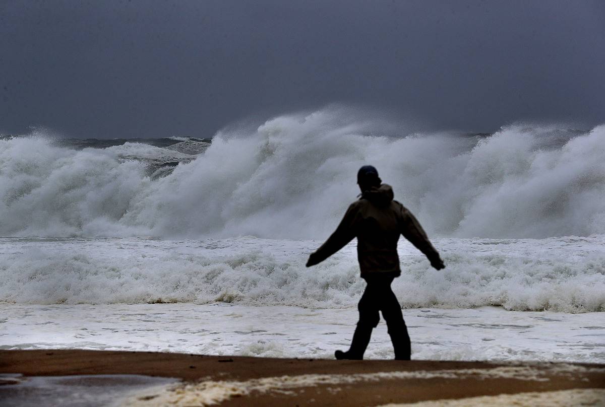A woman walks on the beach next to tall crashing waves.