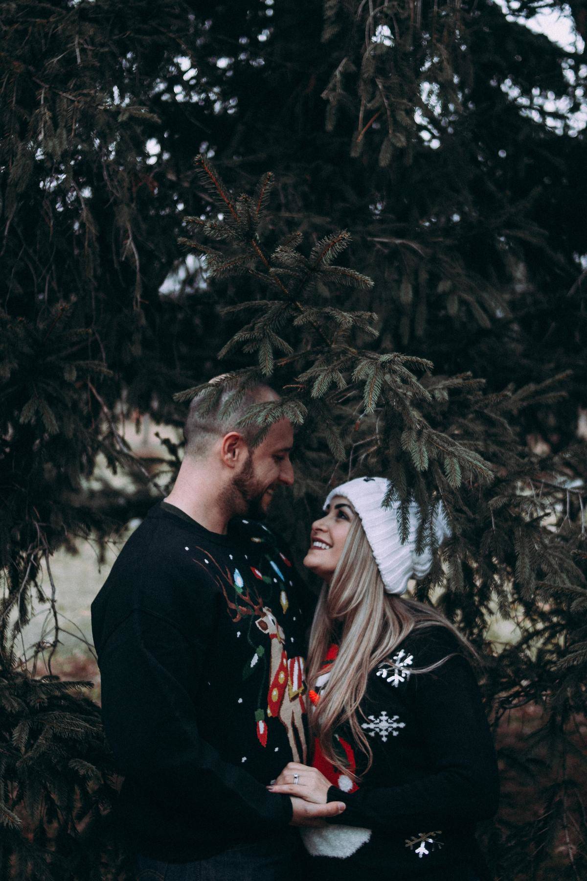 man and woman in christmas sweaters close under pine tree