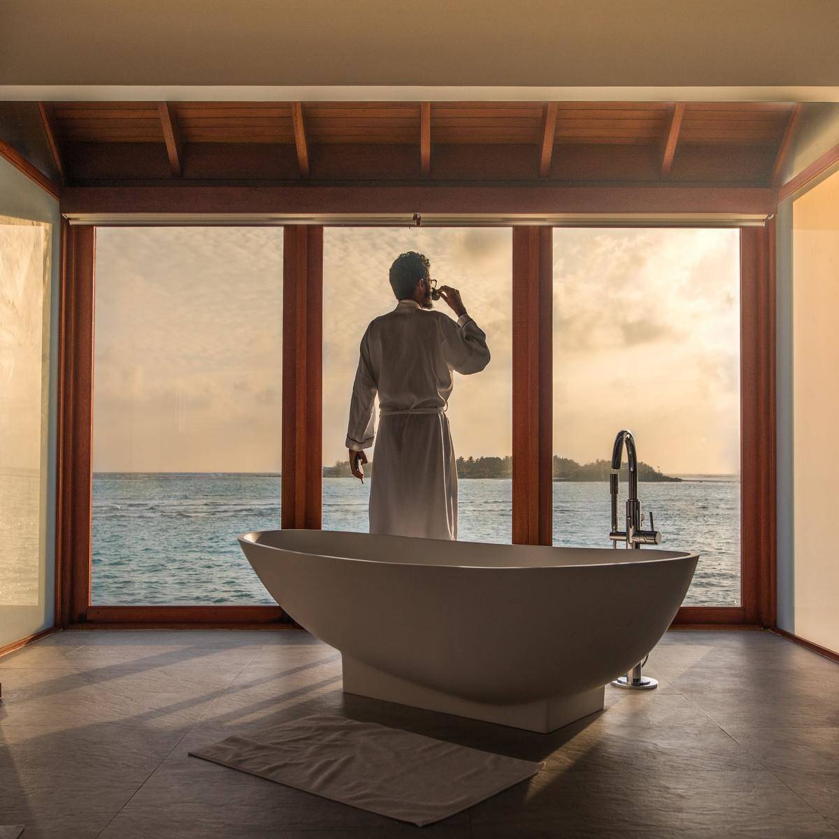 A wealthy man looks out at the ocean while standing in front of his bathtub.