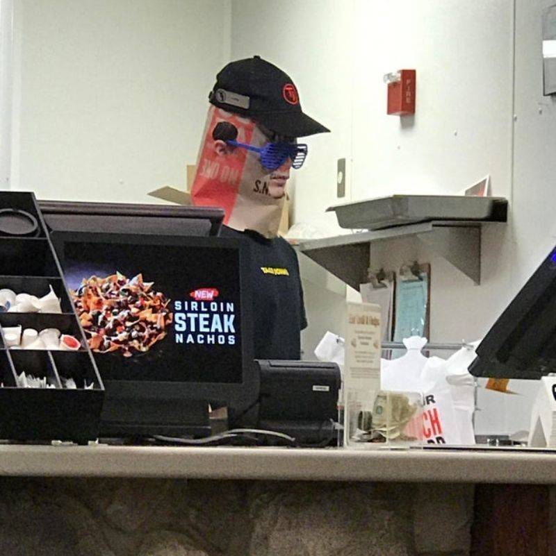 a guy working at a taco place wearing a paper bag on his head with glasses and a hat over it 