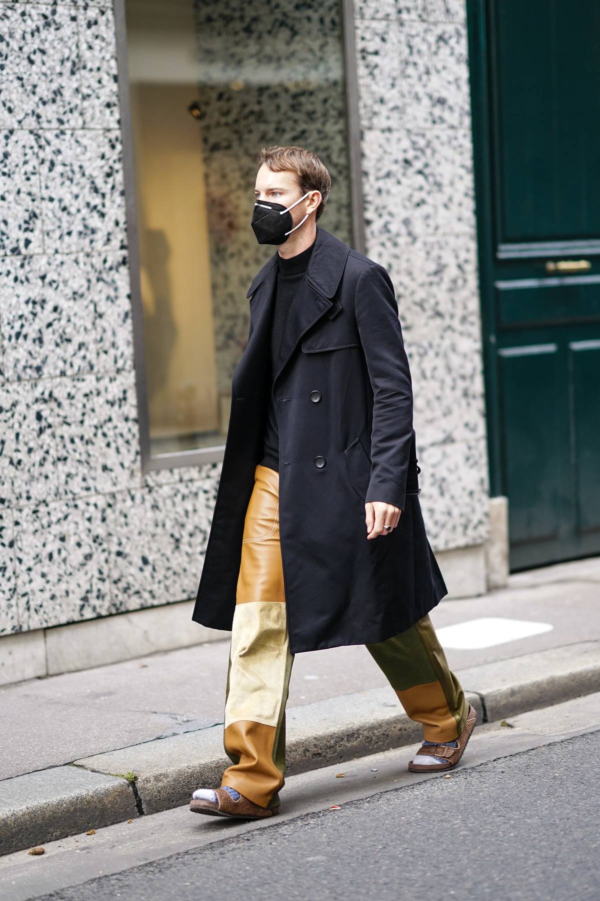 A guest wears a black face mask, a black long coat, a black pullover, brown and beige leather pants, outside Gabriela Hearst, during Paris Fashion Week -