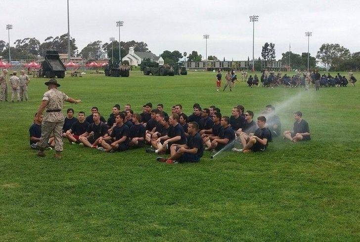 drill sergeant lecturing group of kids sitting outdoors while one is getting hit directly by a sprinkler
