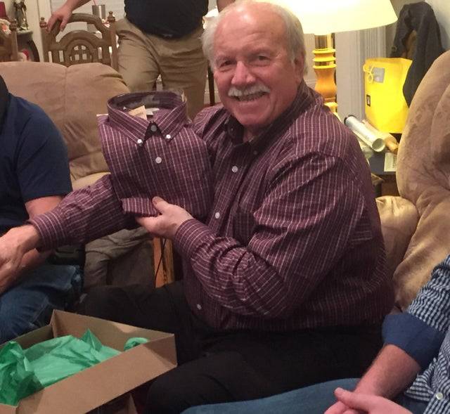  a grandfather smiled for the camera as he holds up his christmas present - which is the exact same shirt that he is wearing 