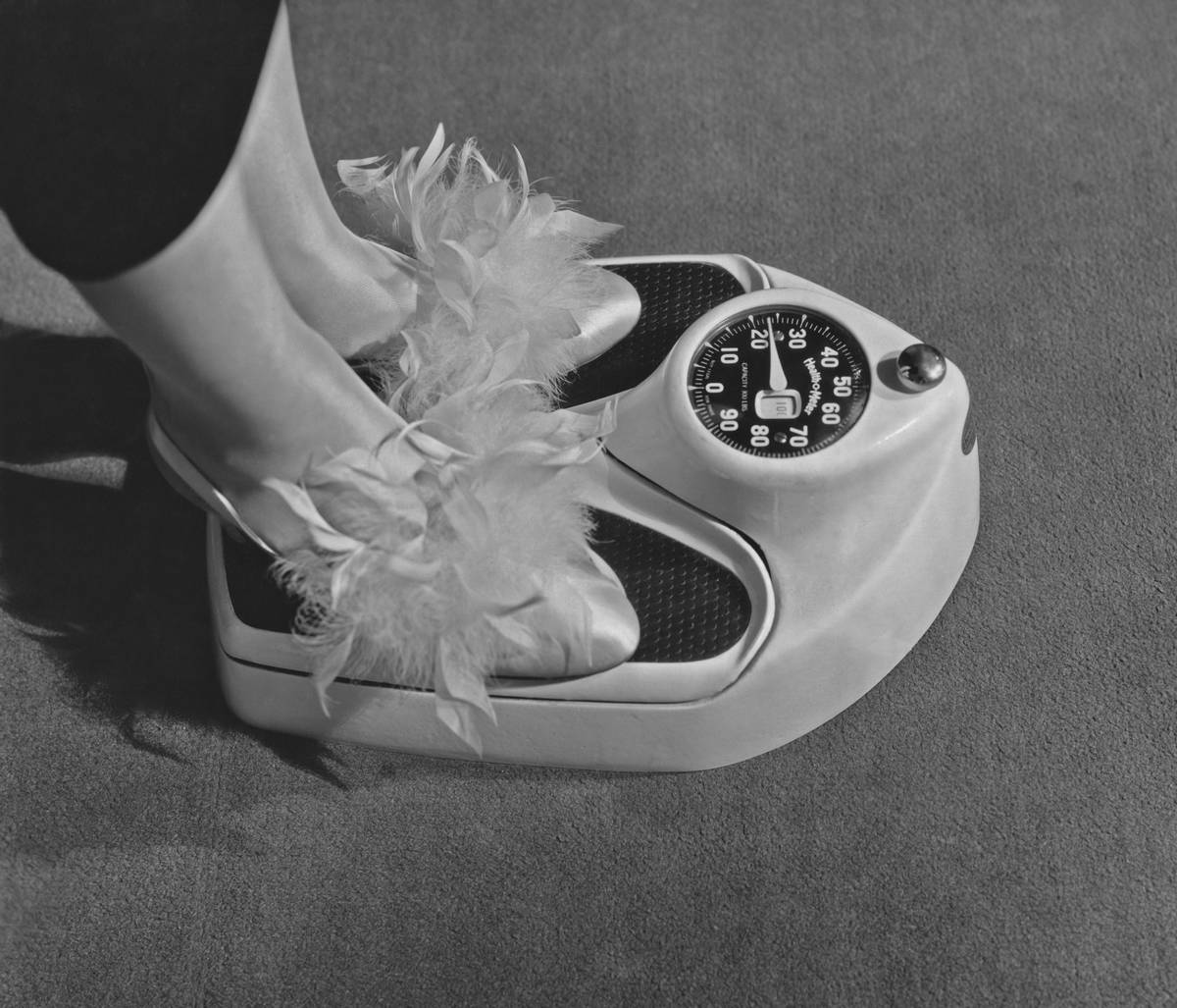 A woman with slippers on steps on a scale to weigh herself.