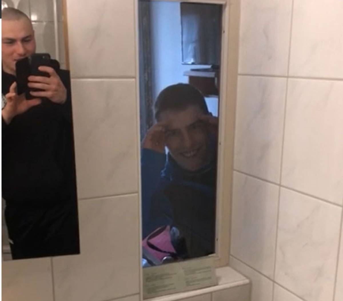 man photographs window from bathroom into bedroom with friend looking through it