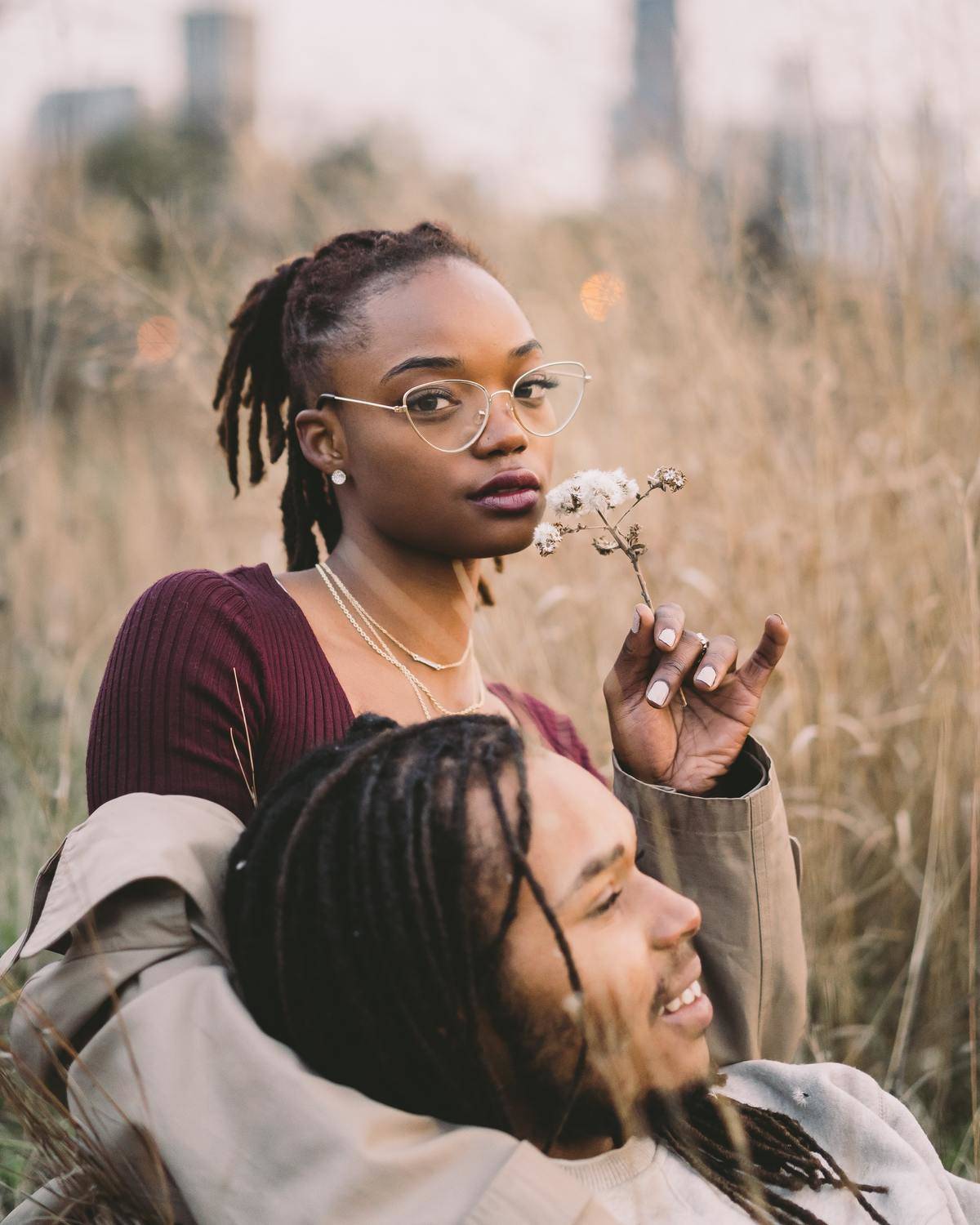 woman sniffing flower in a field while man lays on her