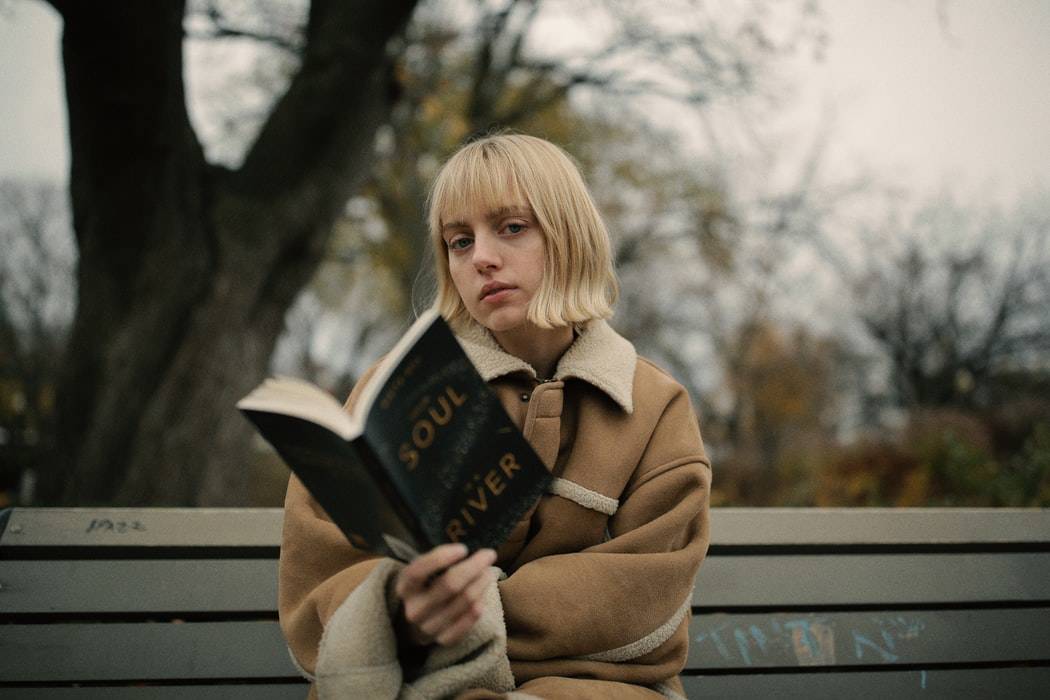 woman reading a book on a park bench as she looks up at the camera 