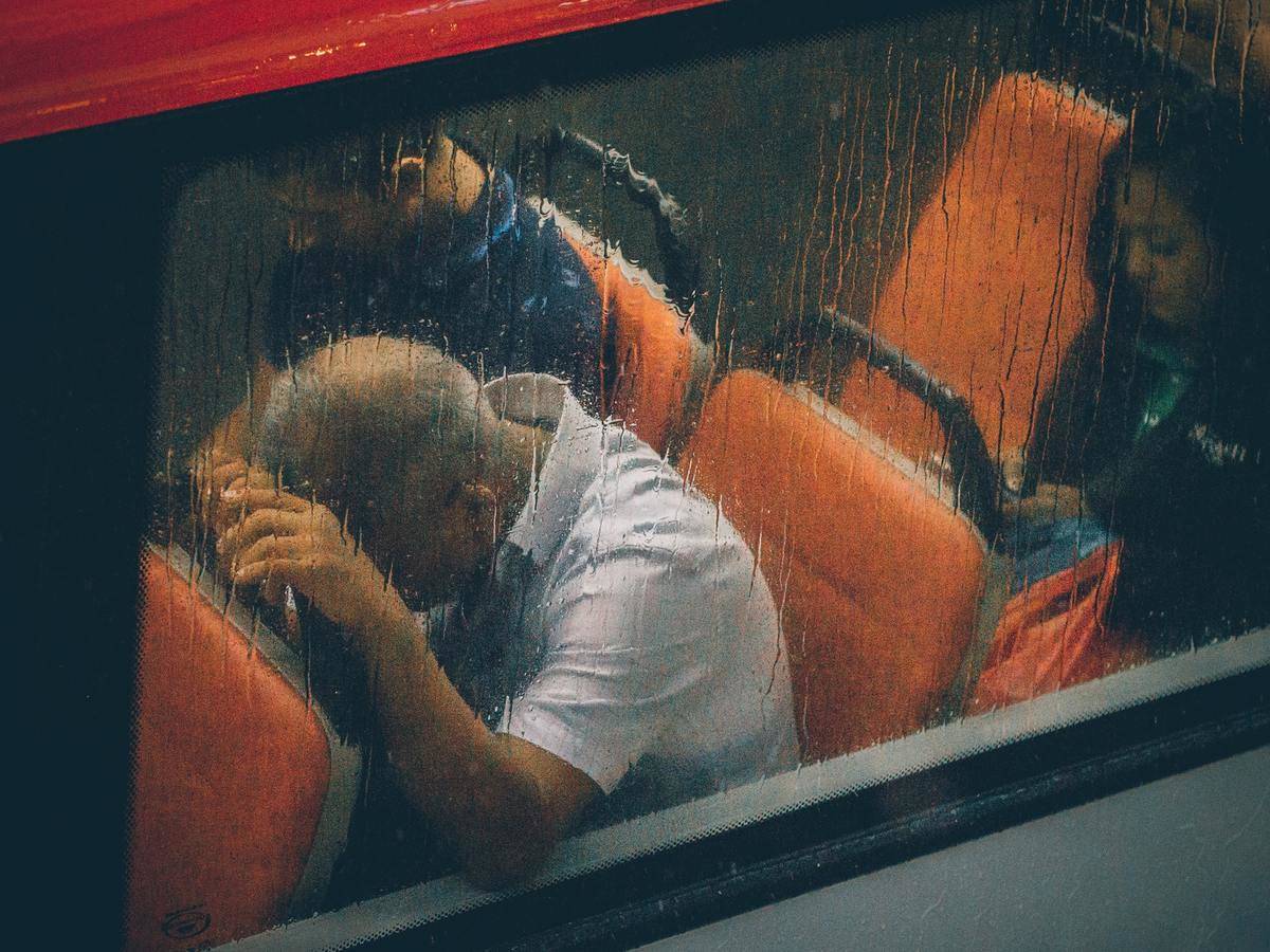 A man presses his head on the back of a bus seat as rain pours on the window.