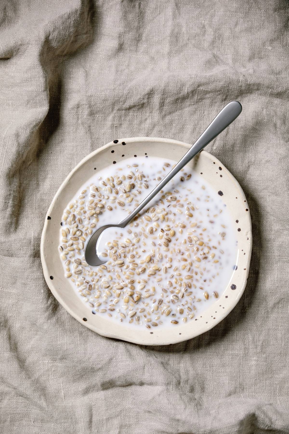 Healthy breakfast organic sprouted wheat with milk in spotted ceramic plate. with spoon over grey linen cloth as background. Flat lay. space
