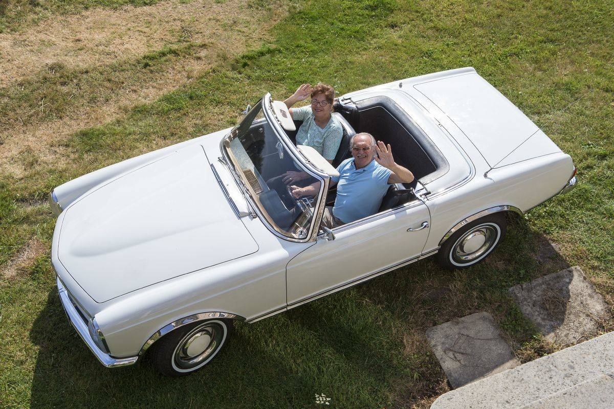 A senior couple waves to the camera from a convertible. 