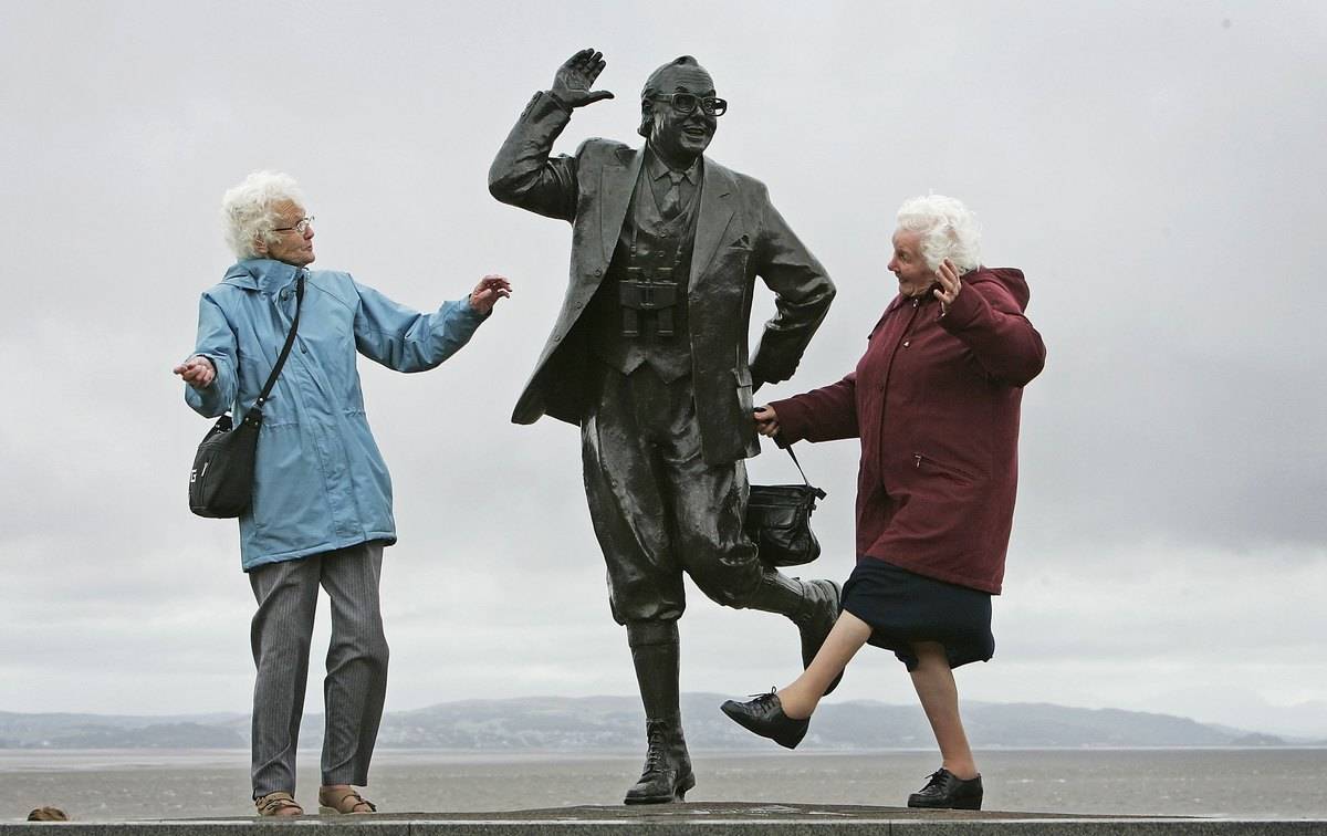 Two senior women perform a famous comedic dance next to a statue of Eric Morcambe.