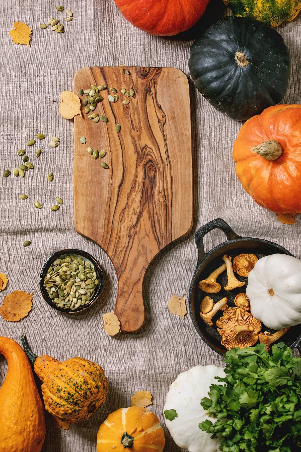 Variety of colorful pumpkins, edible and decorative, with autumn leaves, oregano greens, chanterelles mushrooms, pumpkin seeds and empty wooden cutting board over grey linen cloth. Flat lay, space.