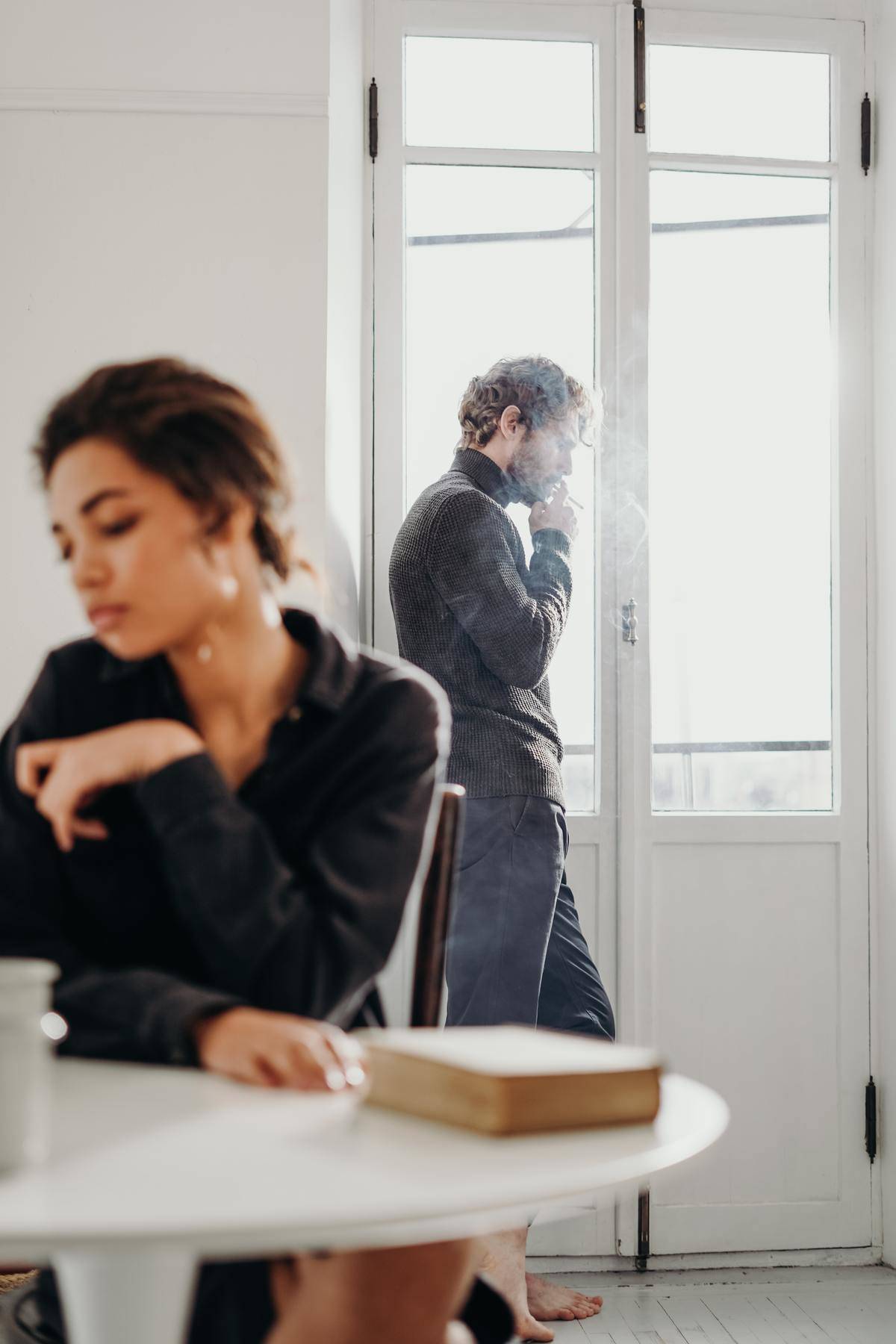 Man stands at balcony window smoking and woman sits at table
