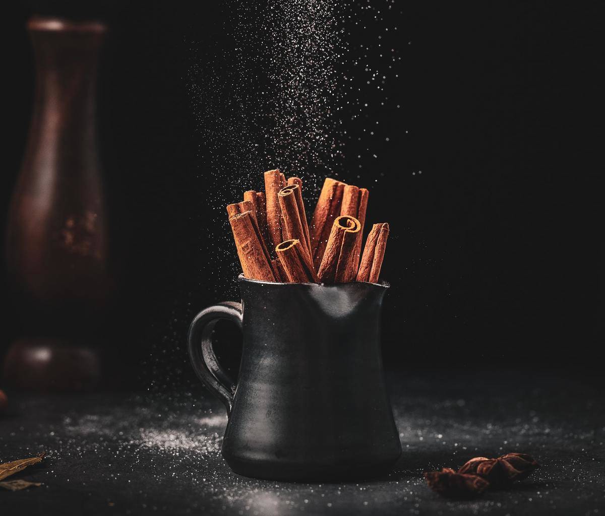 Cinnamon sticks are in a black mug, and powdered cinnamon is sprinkled on it.