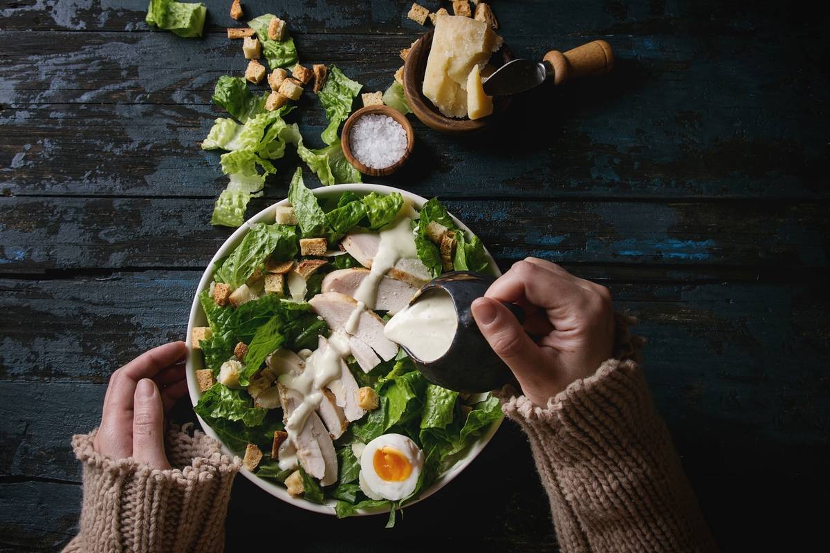 A woman pours Caesar dressing on a salad.