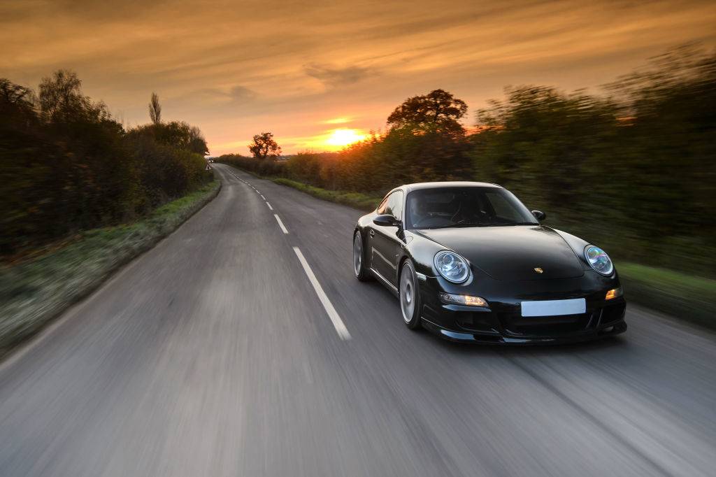 A modified Porsche 997 Carrera S sports car driving on a rural road with a sunset visible in the background